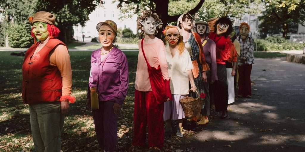 Gruppe von Menschen mit kunstvollen Masken steht in einer Reihe auf einem Parkweg, umgeben von Bäumen und Herbstlaub. Unterschiedliche Kleidung, teils bunt, teils neutral. Eine Person hält einen Korb. Die Szene wirkt ruhig und geheimnisvoll.